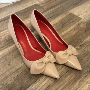 Beige pumps med rosett - Säljer ett par eleganta beige pumps med spetsig tå och en söt rosett framtill. Skorna har en röd sula och klack, vilket ger dem en lyxig touch. Perfekta för festliga tillfällen!