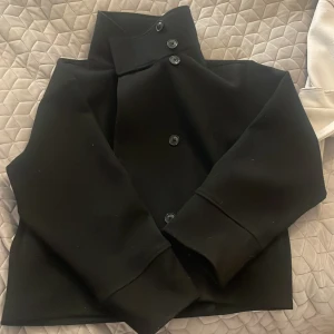 Svart jacka med hög krage - Säljer denna Gina kappa, svart med hög krage och knappar framtill. TRASIG I ENA FICKAN DÄRAV PRISET