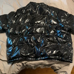 Svart glansig pufferjacka - Säljer en svart glansig pufferjacka som är perfekt för kalla dagar. Jackan har en quiltad design och är långärmad med dragkedja framtill. Den är både stilren och varm, vilket gör den idealisk för vinter och höst. Passformen är normal och materialet är syntetiskt, vilket ger en cool look.