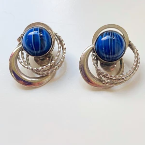 Vintage örhängen med blå sten - Säljer ett par unika vintage clipsörhängen med en stor blå sten i mitten. Troligen glas. De har en cool design med tvinnade silverringar runt stenen. Perfekta för att ge en retro touch till din outfit! Okänd metall 