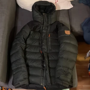 Svart dunjacka från Fjällräven - Säljer en svart dunjacka från Fjällräven i mycket bra skick. Jackan har en hög krage och huva för extra värme. Den är långärmad med dragkedja framtill och har Fjällrävens logga på ärmen. Perfekt för kalla vinterdagar!