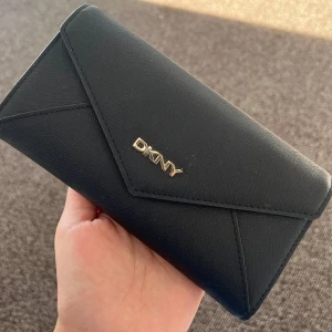 Svart plånbok från DKNY - Snygg och stilren svart plånbok från DKNY i mycket bra skick. Den har flera kortfack och en genomskinlig ficka för ID-kort. Perfekt för att hålla ordning på dina kort och kontanter. Enkelt och elegant design med en liten logotyp i guld på framsidan.
