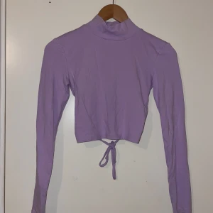 Lila topp med öppen rygg - Säljer en superfin lila topp med lång ärm och hög krage. Toppen har en öppen rygg med knytning, vilket ger en snygg detalj. Perfekt för både vardag och fest! 💜