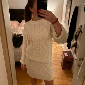 Vit stickat sett med kjol och tröja - Superfin vit stickad klänning med snyggt flätmönster. Den är perfekt för kyligare dagar och har en mysig känsla. Klänningen är kort och har långa ärmar, vilket gör den både stilren och bekväm. Passar perfekt till höst och vinter!