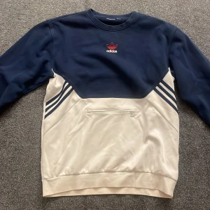 Blå och vit tröja från Adidas - Säljer en snygg blå och vit tröja från Adidas med det klassiska logotypen på bröstet. Tröjan har långa ärmar och en praktisk ficka framtill. Då lappen inte finns med så säljer jag den endast för 250kr! 