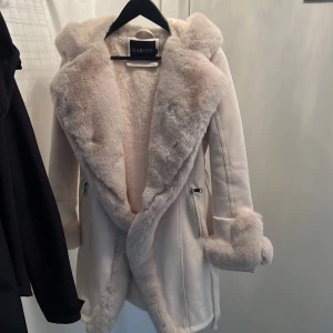 Vit pälsjacka från Garoff - Superfin vit pälsjacka från Garoff med fluffig fuskpäls och huva. Den har långa ärmar och ett bälte i midjan för en snygg passform. Perfekt för kalla vinterdagar när du vill hålla dig varm och stilren. Jackan har också praktiska fickor med dragkedja. Original pris 2500 kr.