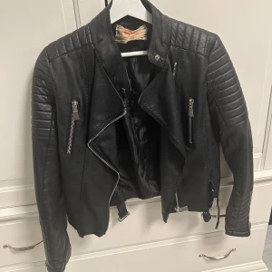 Svart skinnjacka med dragkedjor - Säljer en snygg svart skinnjacka i mycket bra skick. Jackan har coola dragkedjedetaljer och quiltade axlar som ger en edgy look. Perfekt för höst och vår, och funkar lika bra till fest som till vardags. Passformen är normal och den har långa ärmar. Ett måste i garderoben för den som gillar rockig stil! Storlek 40, men ganska liten i storleken som motbevisar S