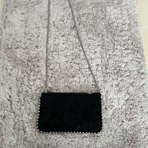 Svart axelväska med kedja - Snygg svart axelväska i syntetmaterial med en cool kedja som axelrem. Väskan har en dragkedja upptill och är perfekt för en utekväll eller fest. Den är liten och smidig, men rymmer det viktigaste. Perfekt för att ge din outfit en edgy touch!. Den är lite sönder på dragskedian därför säljer jag den mindre 