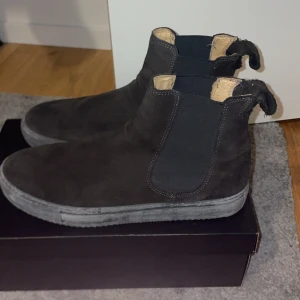 Svarta mocka Chelsea boots - Säljer ett par snygga svarta Chelsea boots i mocka. De har en klassisk design med elastiska paneler på sidorna för enkel på- och avtagning. Perfekta för både höst och vår, och de ger en stilren touch till vilken outfit som helst. Skorna är i bra skick och redo för nya äventyr!
