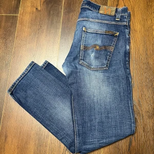 Blå jeans från Nudie Jeans - Säljer ett par snygga blå jeans från Nudie Jeans i bra skick. Storlek W32, L30. De har en klassisk femficksdesign med orange sömmar och en knappgylf. Perfekta för vardagsbruk och passar till det mesta. Obs ett litet hål i bakre ficka. 