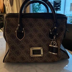 Guess väska - Vintage handväska från Guess, jätte fin men har använts därav är dragkedjan lite trasig (möjlig att fixa) samt handtagen har slitage. I övrigt riktigt snygg och elegant väska!   Guld detaljer, berlocker och många innefickor.