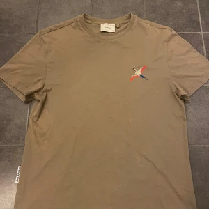  t-shirt från Axel Arigato - Säljer en snygg beige t-shirt från Axel Arigato i mycket bra skick. Den har en cool broderad fågel i rött, blått och grått på bröstet. Perfekt för en avslappnad stil! 🕊️Stolek m herr. Bra skick använd några gånger.  nypris 1099 köpt på nk i Stockholm. Priset är ganska fast. Går att pruta. Bara att skriva ifall ni har frågor! 