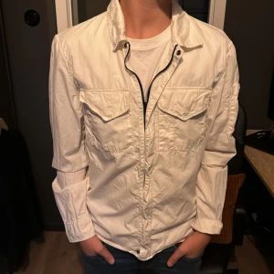 Vit jacka från C.P. Company - Säljer en snygg vit jacka från C.P. Company i mycket bra skick. Den har en stilren design med dragkedja och två bröstfickor med lock. Perfekt för vår och höst! Jackan är långärmad och har en cool detalj på ärmen. Passar perfekt till både vardag och fest.