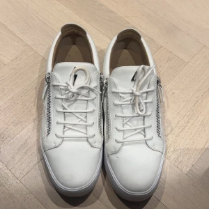 Vita sneakers med dragkedja (Zanotti giuseppe) - Säljer ett par stilrena vita sneakers i skinn med snörning och dragkedja på sidan. Perfekta för både vardag och fest! De är i mycket bra skick och passar till alla säsonger. Skorna har en klassisk design med en modern twist tack vare dragkedjan. Storlek 44 men passar också 45 bra.