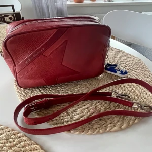 Röd axelväska i skinn - Säljer nu min röda väska från Golden Goose inköpt år 2021. Den har tyvärr en liten defekt på bild nr 3 som inte syns särskilt mycket och man kan säkert fixa det, därav drar jag ner priset lite. Den är annars i väldigt bra skick och väldigt rymlig🤗❤️Nypris på väskan ungefär 6000kr!