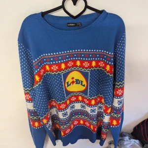 Blå jultröja från Livergy - Blå jultröja från Livergy med Lidl-motiv. Tröjan har ett färgglatt mönster med snöflingor och granar i rött, gult och vitt. Perfekt för julfesten eller som en kul present! Den är långärmad och gjord i 100% acrylic 