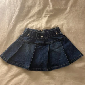 Mörkblå minikjol med dragkedja - Trendig mörkblå jeanskjol (minikjol) med dragkedja i baksidan och dekorativa knappar. Kjolen har en kort och utsvängd design med veckade detaljer som ger en snygg look. 