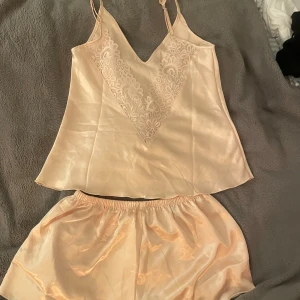 Champagne-färgad pyjamasset med spets - Säljer ett jättefint satinpyjamas-set med v-ringad spetsdetalj till ryggen. Axelbanden är justerbara och materialet på pyjamassetet är skönt och tunnt. Aldrig använd!!