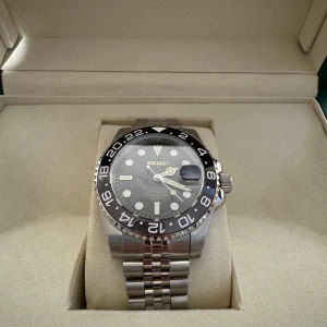 Seiko GMT Bruce Wayne - Säljer en stilren Seiko-klocka med svart urtavla och silverfärgad boett. Klockan har en tydlig 24-timmarsmarkering på ringen och ett robust armband i silver. Perfekt för den som söker en kombination av funktion och stil.