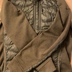 Olivgrön jacka från Peak Performance - Säljer en olivgrön jacka från Peak Performance med quiltade detaljer och dragkedja. Jackan har en stilren design med långärmade ärmar och en praktisk bröstficka med dragkedja. Perfekt för kyligare dagar.