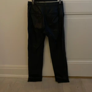 Svarta skinnbyxor - Snygga svarta skinnbyxor med en tight passform. Perfekta för en stilren look. Byxorna har en hög midja och är tillverkade i ett glansigt material som ger en cool känsla. Innesömnen på byxans ena sida har gått upp vilket gör att tyg går ut lite längst ner på byxan, som bilden visar. Storlek 34