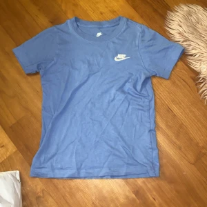 Blå t-shirt från Nike - Säljer en blå t-shirt från Nike med korta ärmar och klassisk rund halsringning. T-shirten har en vit Nike-logga broderad på bröstet. Perfekt för en sportig look eller avslappnad stil. Passar bra till  träningsbyxor.