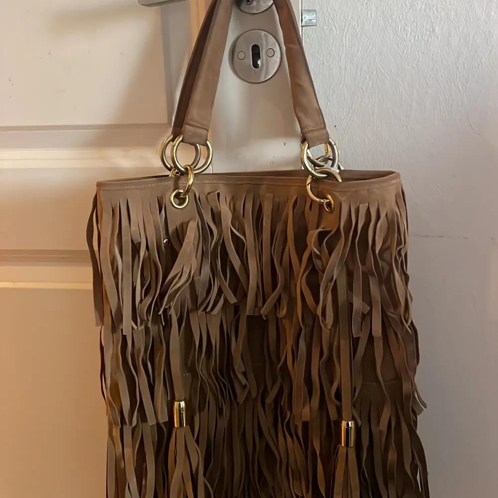 Säljer en snygg brun axelväska med fransar. Väskan har två handtag och är dekorerad med guldfärgade detaljer. Insidan är rymlig med flera fack för enkel organisering. Perfekt för att ge din outfit en bohemisk touch. Äkta Mocca. Laukut & Käsilaukut.