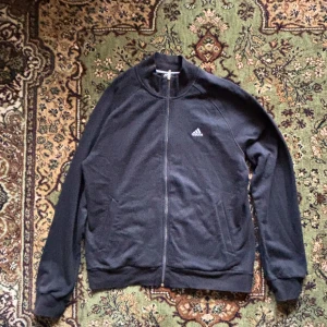 Svart träningsjacka från Adidas - Säljer en svart träningsjacka från Adidas med dragkedja och logga på bröstet. Jackan är långärmad och tillverkad i ett bekvämt material, perfekt för träning eller vardagsbruk.
