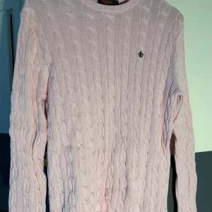 Ljusrosa stickad tröja från Morris - Säljer en ljusrosa stickad tröja från Morris med kabelstickat mönster. Tröjan har långa ärmar och en liten broderad logotyp på bröstet. 