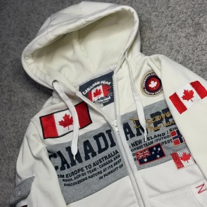 Canadian Peak Hoodie - Säljer en vit huvtröja köpt från Zalando, med Canadian Peak detaljer och flera flaggor som textdetaljer. Tröjan har en dragkedja framtill och en stor huva med snörning. Perfekt för en avslappnad stil med en touch av äventyr.