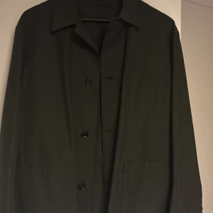 Svart rock från Massimo Dutti - Elegant svart rock från Massimo Dutti med klassisk krage och knäppning framtill. Rocken har långa ärmar och två stora fickor framtill. Perfekt för en stilren look.