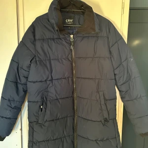 Mörkblå dunjacka från CRW - Säljer en mörkblå dunjacka från CRW med dragkedja framtill och två praktiska fickor. Jackan har en quiltad design och långa ärmar, perfekt för kyliga dagar. Den är både stilren och funktionell.