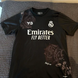 Svart Real Madrid fotbollströja - Snygg svart Real Madrid tröja med Y-3 och Emirates loggor. Unikt ros-mönster på framsidan och baksidan. Perfekt för fans som vill ha något speciellt i garderoben. Lite flisig på ryggen men det är inget som syns.