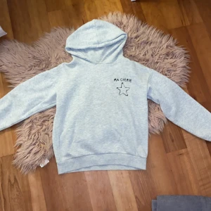 Grå hoodie från Gina Tricot - Säljer en grå hoodie från Gina Tricot med texten 'Ma Chérie' och en stjärna tryckt på både fram- och baksidan. Tröjan har långa ärmar och en bekväm huva. Perfekt för en avslappnad stil. 🩶