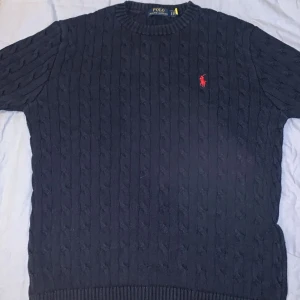 Mörkblå stickad tröja från Polo Ralph Lauren - Säljer en snygg mörkblå stickad tröja från Polo Ralph Lauren med det klassiska röda logotypbroderiet på bröstet. Tröjan har en ribbad rund hals och ribbade muddar vid ärmslut och nederkant. Perfekt för kyligare dagar!