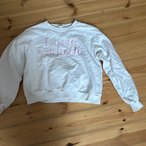 Vit sweatshirt från Lindex med text - Säljer en vit sweatshirt från Lindex med texten 'La vie est belle' i rosa på framsidan. Tröjan har långa ärmar och en rund halsringning. Perfekt för en avslappnad stil. Strl 14/152 barn 