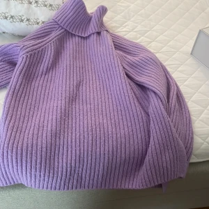 Lila stickad tröja från Gina Tricot - Mysig lila stickad tröja från Gina Tricot med ribbad design och hög krage. Perfekt för kyliga dagar och ger en stilren look. Passar bra till jeans eller kjol för en avslappnad stil. Väldig mysig oversize tröja med slits på sidorna 