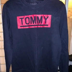 Svart tröja från Tommy Hilfiger - Säljer en svart tröja från Tommy Hilfiger med röd text på bröstet. Tröjan har långa ärmar och en rund halsringning. Perfekt för en casual look. Litet märke vid Y som möjligt går bort efter en tvätt. Hör av dig vid intresse! 