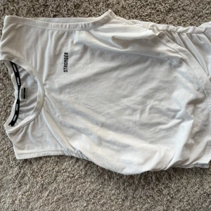 Vit ärmlös topp från Stronger - Säljer en vit ärmlös topp från Stronger i storlek S. Perfekt för träning eller en avslappnad dag. Toppen har en enkel design med märkets logga tryckt på framsidan och längs insidan av kragen.