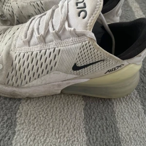 Nike Air Max 270 i vitt - Säljer ett par vita Nike Air Max 270 sneakers med svart Nike-logga och synlig Air-enhet i hälen. Skorna har snörning och en modern design med ventilerande material.