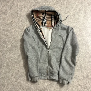 Snygg gråa burberry zip hoodie  - Säljer en snygg stilig gråa burberry, helt okej skick finns en liten skada som märks inte när man har på sig den d blandas in med färgen. Storlek L men passar som M