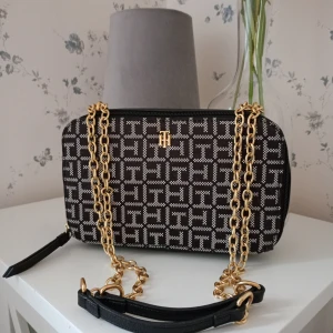 Svart och vit axelväska med kedjedetaljer - Snygg axelväska i svart och vitt med ett geometriskt mönster. Väskan har guldfärgade kedjedetaljer och en dragkedja för säker förvaring. Perfekt för att ge din outfit en elegant touch.