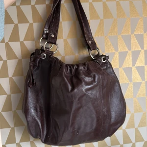 Brun axelväska i skinn - Säljer en snygg brun axelväska i skinn med praktiska handtag och en dragkedja på insidan. Väskan har en stilren design och är perfekt för att bära med sig det nödvändigaste. Den har även en knappstängning upptill för extra säkerhet.