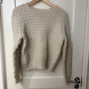Beige stickad tröja från Vero Moda - Mysig beige stickad tröja från Vero Moda. Perfekt för kyliga dagar med sin mjuka och fluffiga textur. Tröjan har långa ärmar och en rund halsringning, vilket gör den både stilren och bekväm.