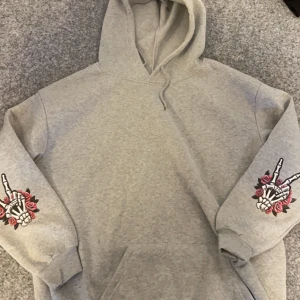 Grå hoodie med tryck - Säljer en grå hoodie med coolt tryck av en skelettfigur och texten 'Expensive Talks Back' på ryggen. Ärmarna har tryck av skelett händer med rosor. Perfekt för en avslappnad stil.