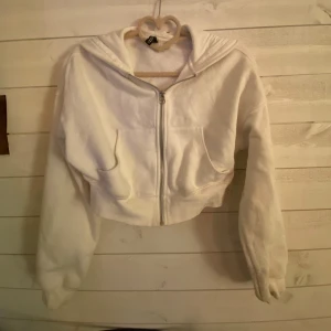 Vit croppad hoodie från H&M - Säljer en vit croppad hoodie från H&M med dragkedja och huva. Den har långa ärmar och är perfekt för en avslappnad stil. Hoodie är i en mjuk och bekväm bomullsmix.