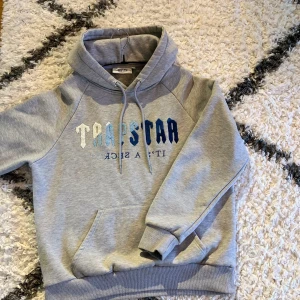 Äkta Trapstar hoodie - Säljer en grå hoodie från Trapstar med broderad text i olika blå nyanser på framsidan. Hoodien har en klassisk design med dragsko i huvan och en stor magficka. Perfekt för en avslappnad stil.               Äkta! Säljer på grund utav att den är för kort för mig. OBS… Snodarna på luvan är ej kvar!