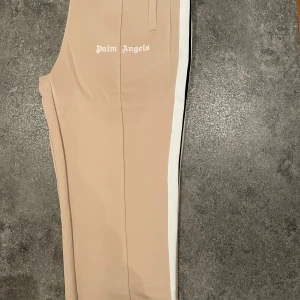 Beige byxor från Palm Angels - Snygga beige byxor från Palm Angels med vit sidoremsa. Perfekta för en stilren look. Byxorna har en normal passform och är tillverkade i ett bekvämt material.