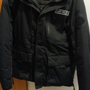 Svart jacka från Black Squad - Säljer en svart jacka från Black Squad med coola detaljer som broderade märken och text på ryggen. Jackan har dragkedja och flera praktiska fickor. Perfekt för en urban look. Inga skador och använt ett par gånger. Passar också S