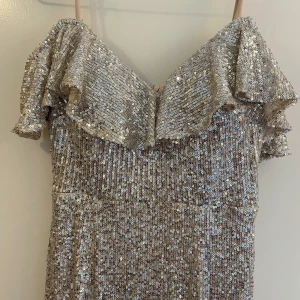 Glittrig långklänning med volangdetaljer - Säljer en fantastisk glittrig långklänning i silver med volangdetaljer vid axlarna. Klänningen har en elegant v-ringning och en vacker slits med inslag av ljusrosa tyg. Perfekt för en glamorös kväll! ✨Köparen betalar frakt. 